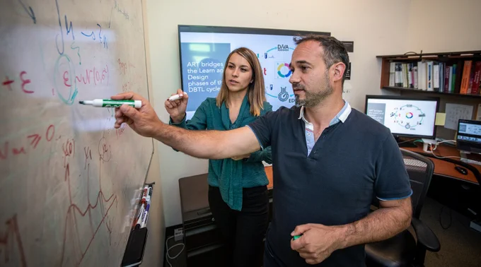  Two researchers stand on the right holding markers toward a large whiteboard. 
