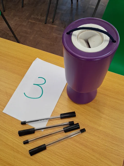 ​A purple collection box, the number 3 written on a large piece of paper, and 4 black biros, all on a table.