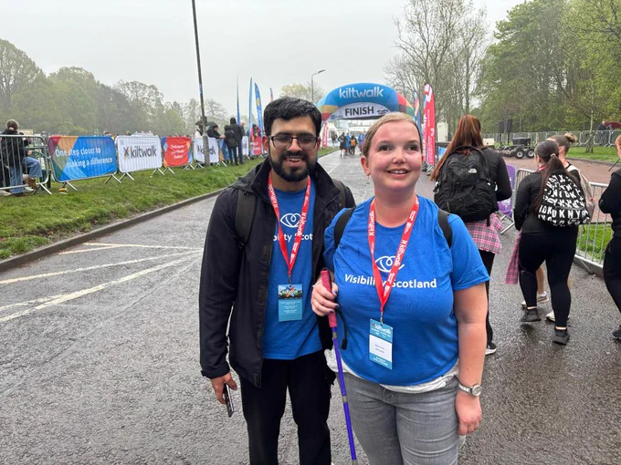 Two people stand together smiling in front of the Glasgow Kiltwalk finish line. They are wearing blue Visibility Scotland t-shirts.