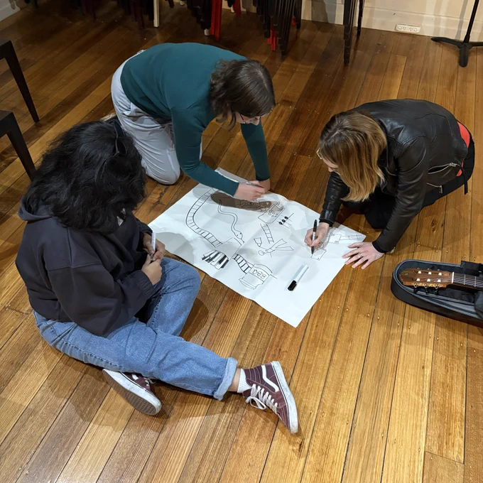 Photo showing three people seated around a large notepad, collaboratively drawing a graphic score. A snake, some ladders and a simple human figure can be seen.