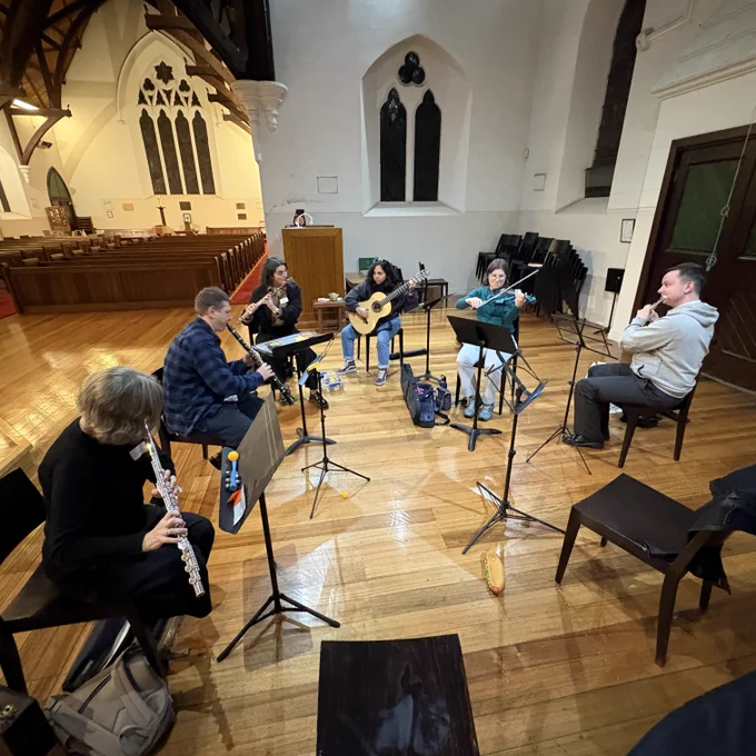 Photo showing five seated musicians playing flutes, clarinet, guitar and violin, inside a church.