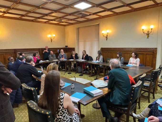 A group of people are seated around a large rectangular table in a formal meeting room with wood-paneled walls and ornate ceiling tiles. Some participants are speaking, while others listen attentively. A sign language interpreter stands at the back of the room. Several individuals have documents and blue booklets in front of them. The room is lit by wall-mounted lamps.
