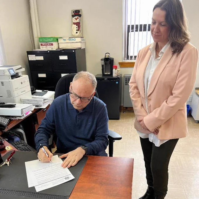 Minister Pike sits at a desk signing a piece of paper. A woman stands beside him.