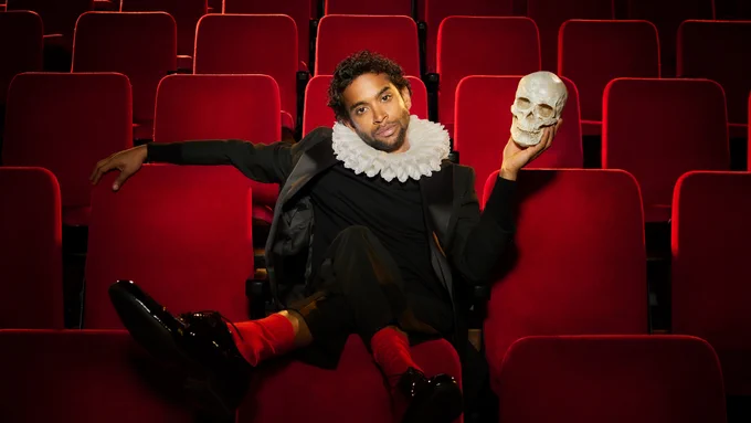 A person in a dark suit and ruffled collar sits dramatically in red theater seats, holding a skull in one hand, evoking a classic Shakespearean pose.