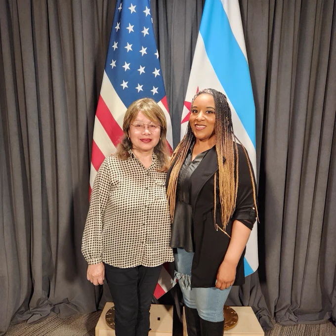 Two individuals posing for a photo in front of the United States and Chicago flags.