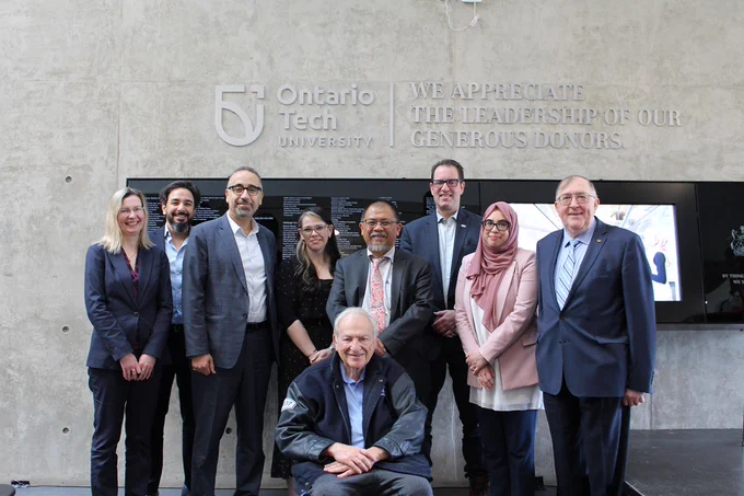 8 people in business attire stand for a photo, one sits infront of them. Behind them is a concrete wall with a sign that says "Ontario Tech University" 