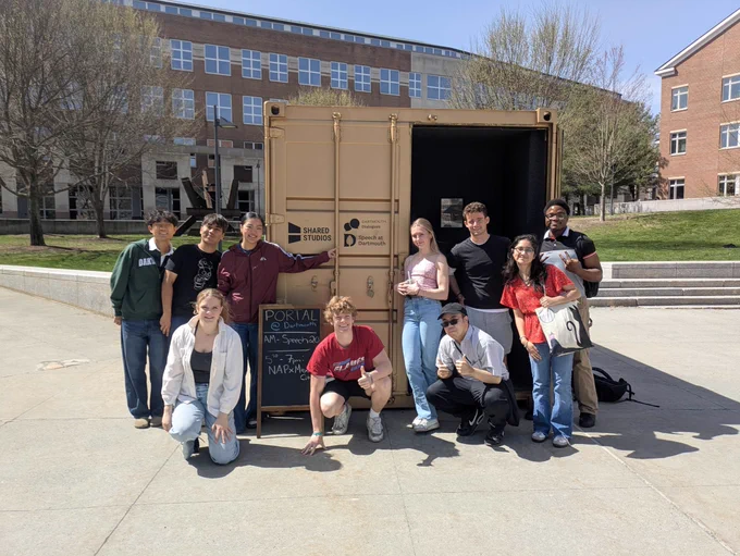 Group of students posing in front of a large shipping container with "Portal @ Dartmouth" written on it, outdoors on a sunny campus setting.