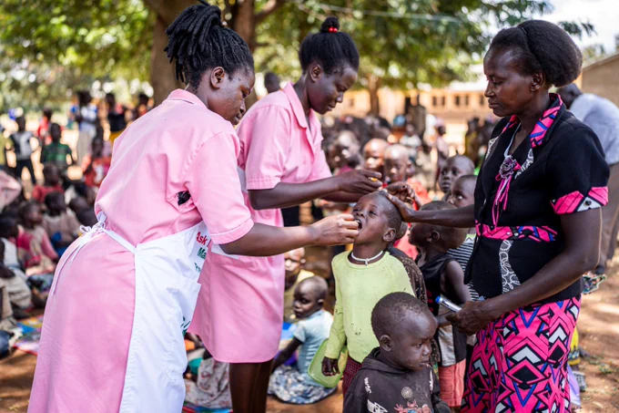 Children line up at an early childhood development center to receive vaccines from two health workers wearing pink.
