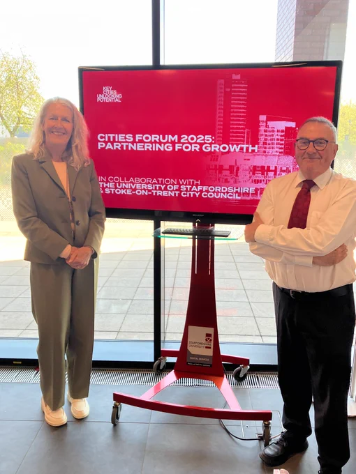 Two smiling attendees standing in front of a display screen showing 'Cities Forum 2025: Partnering for Growth' at Staffordshire University.