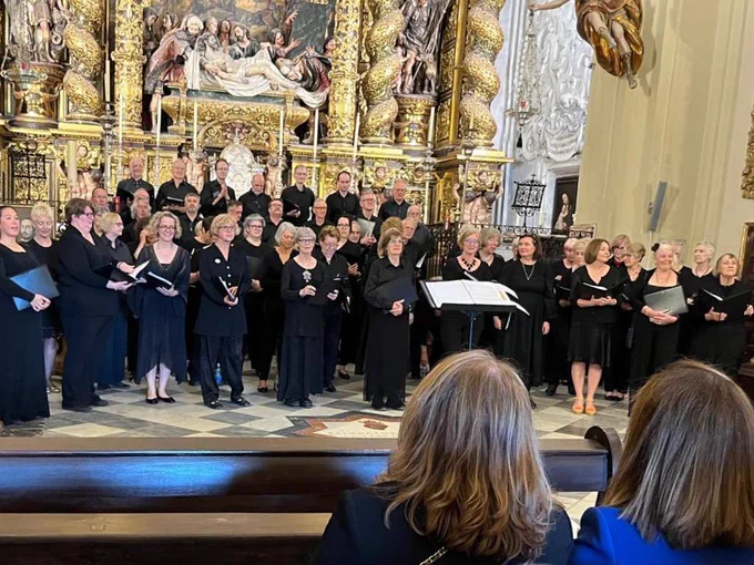 A large choir dressed in black performs in an ornate church, standing in front of an elaborate gilded altarpiece with religious artwork and sculptures. Two audience members are visible in the foreground, seated in pews, watching the performance.