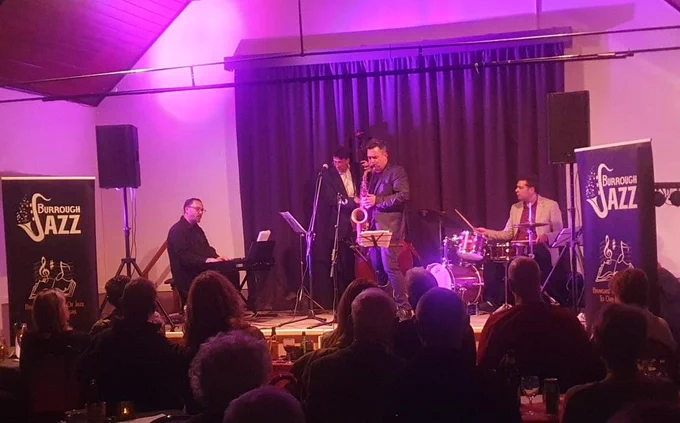 A jazz band plays on stage in a small dimly lit venue. The audience can be seen in the foreground. There is a keyboardist, saxophonist, double bassist, and drummer on stage. There are signs on either side of the stage with the band's details on.