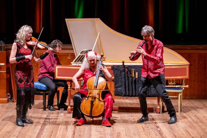 Four musicians perform on a wooden stage in vibrant red and black outfits. A woman plays the violin, a man plays the harpsichord, another woman plays the cello, and a man plays a recorder. Behind them is a grand harpsichord and a curtain lit with warm, colourful stage lights.