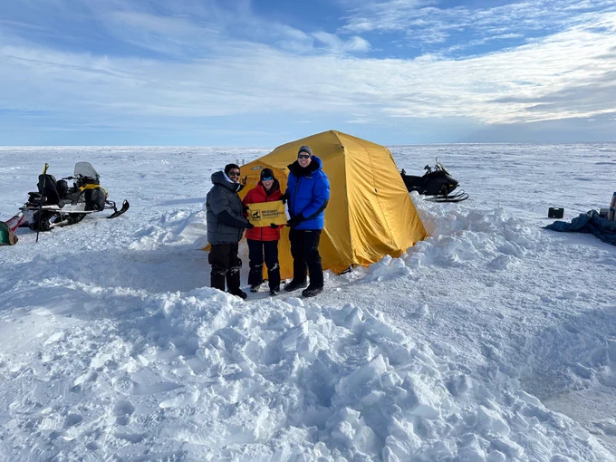 Michigan Tech researchers Stephen Techtmann, Trista Vick-Majors and KM Shafi stand on the ice in the Arctic Circle, north of Utqiagvik, Alaska. They are holding a yellow banner that reads “Michigan Technological University” and standing in front of a yellow lab tent and snowmobiles.

