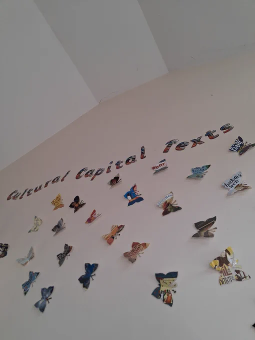 A high wall leading to a ceiling, with over 20 butterflies made out of book covers. Text above reads "Cultural Capital Texts".