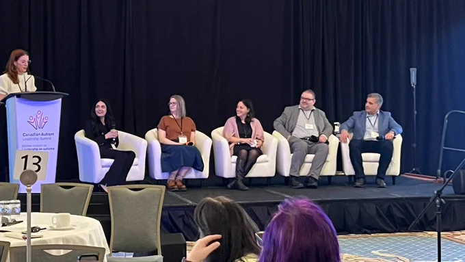 An MC stands at a lectern addressing the Canadian Autism Leadership Summit attendees. Five panelists are seated in a row next to the lectern.