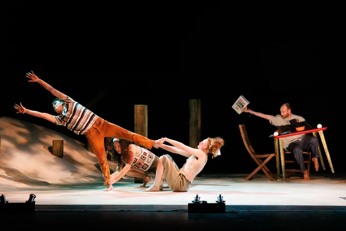 Four performers in a dynamic theater scene; one is balancing on one foot while being held by two others who are on the floor. Another performer sits at a small table.