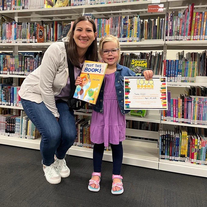A child and caregiver pose for a photo commemorating the completion of 1000 Books Before Kindergarten.