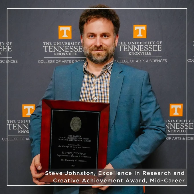Person holding a commemorative plaque stands in front of a University of Tennessee, Knoxville,  College of Arts and Sciences backdrop. The plaque is from the College of Arts & Sciences and recognizes Steve Johnston for winning an Excellence in Research and Creative Achievement Award for his mid-career work.