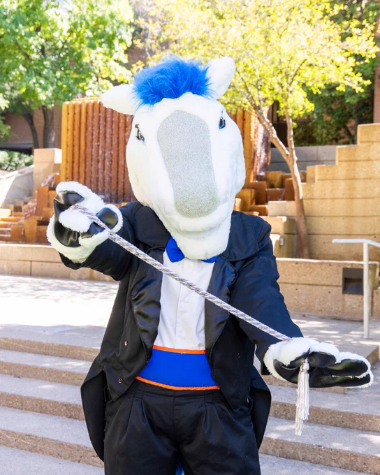 Person in a horse mascot costume, wearing a suit, holding a graduation cord, and standing on stairs outdoors.