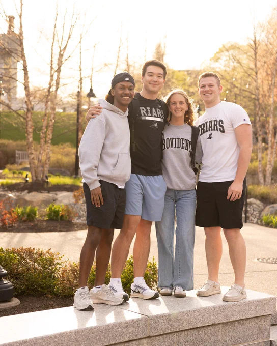 Four individuals are standing closely together, smiling, in a sunny outdoor setting with trees in the background. Two are wearing tops with "Providence College" and "Friars" logos,. They appear to be enjoying a pleasant day outdoors.