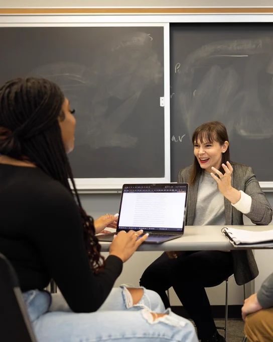 Andrea McDonnell, Ph.D., engages in a discussion with a laptop in a classroom with a blackboard in the background. The student is working on a laptop, and McDonnell is gesturing while speaking.