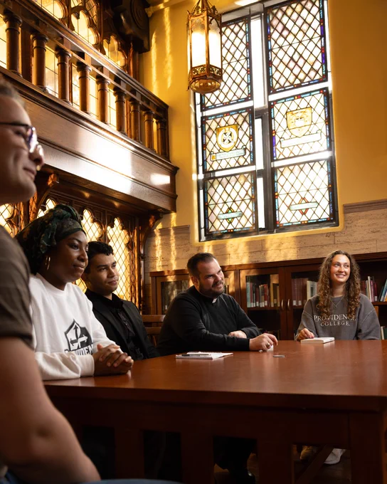 Group of four students and Rev. Justin Brophy, O.P., sit around a wooden table in the Center for Catholic and Dominican Studies with stained glass windows, engaged in a discussion.