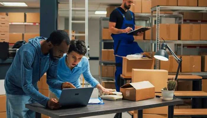 Two men collaborating while working on a laptop at a table in a warehouse filled with cardboard boxes; a third man in blue overalls is checking inventory on a clipboard in the background.