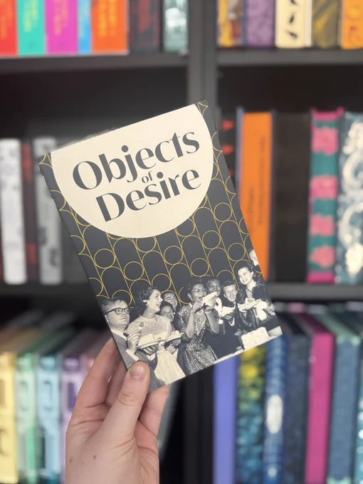 Hand holds "Objects of Desire" book with a black and white cover showing people celebrating. Colorful books on shelves in the background.