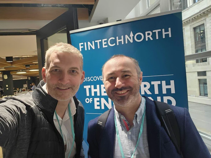 Two smiling men take a selfie in front of a "Fintech North" banner at an event.