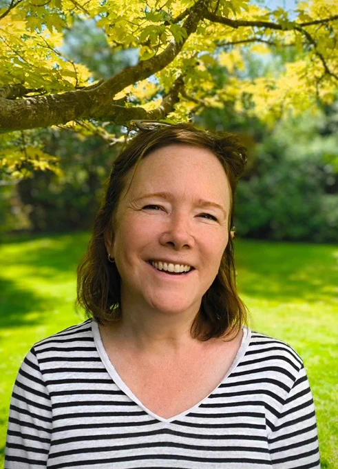 Headshot photograph of Amanda. She is outside on a bright day with trees and a field in the background. She is smiling warmly at the camera. Her glasses are resting on her head. She is wearing a Breton top.