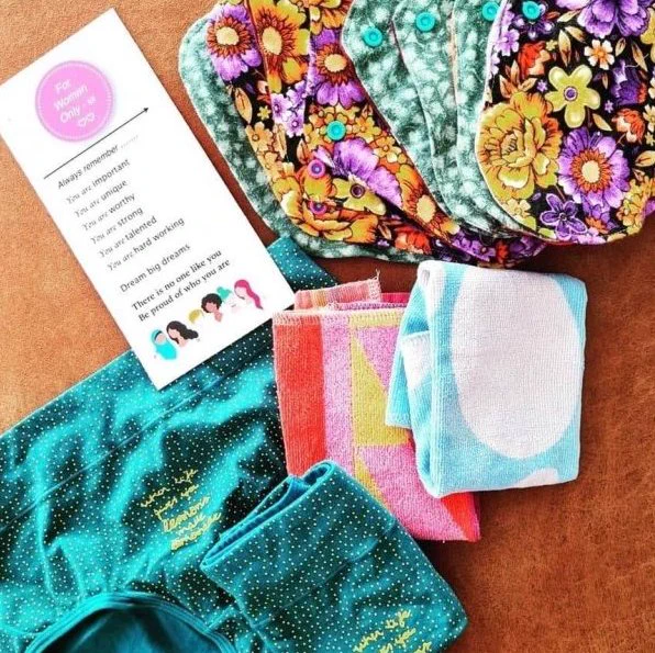 A set of reusable menstrual hygiene products displayed on a brown surface. The set includes colourful patterned cloth pads, a teal-coloured underwear set with gold dotted patterns, small towels, and an inspirational card that reads, “For Women Only” and encourages women to be proud of who they are. The overall display promotes dignity and empowerment through sustainable period care.