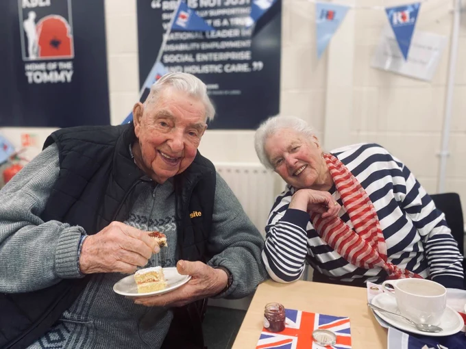 An older couple smile during an afternoon tea to commemorate the 80th anniversary of Victory in Europe Day. They have cake and Union Jack serviettes 