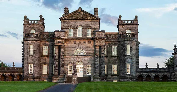 Front view of Seaton Delaval Hall, featuring ornate stone architecture with symmetrical facades and a central arched window, set against a twilight sky.