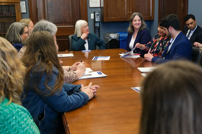 Senator Murray is sitting at the end of a table with her hand resting on her chin. She is looking to the right and smiling. 10 other people are sitting at the table.