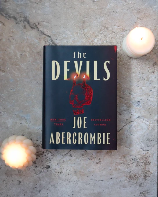 The Devils by Joe Abercrombie is on a marble flatlay with two lit white candles 