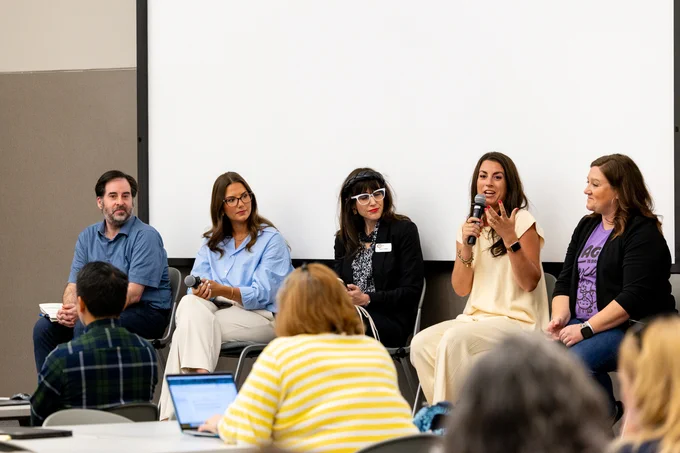 Five panelists, including one holding a microphone, are seated at the front of a conference room while engaging with an audience during a discussion session.