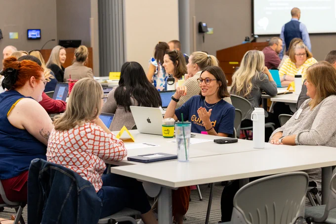 Participants engaging in a group discussion at a workshop with laptops and notebooks on desks, in a classroom setting.