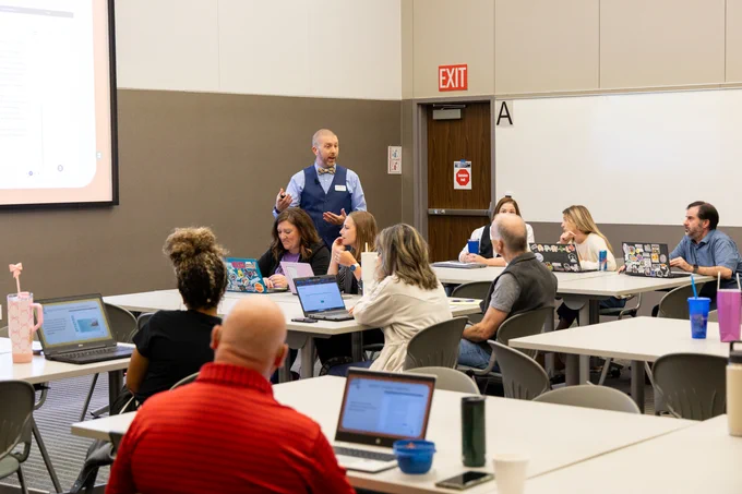 A group of adults attentively participating in a workshop. One person is standing and speaking at the front near a digital presentation screen, while others are seated at tables with laptops.