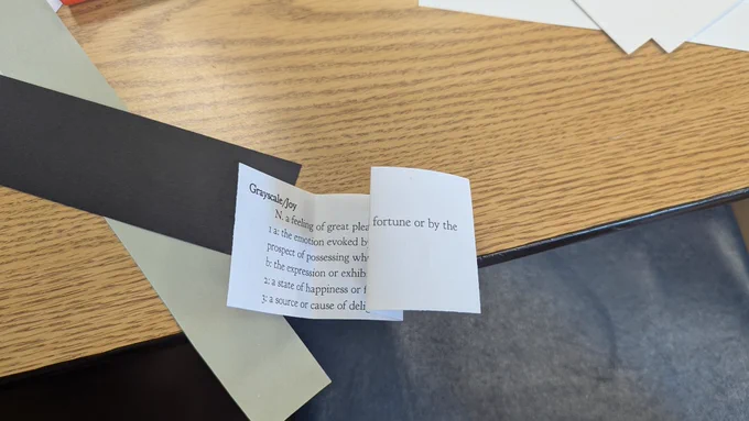 Strips of black and grey paper with a slip of paper that says "Greyscale/Joy"