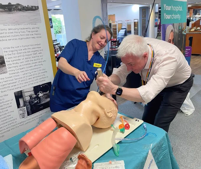 Two healthcare professionals, one in blue scrubs and the other in a white shirt and tie, are practicing intubation on a training mannequin in a hospital setting.