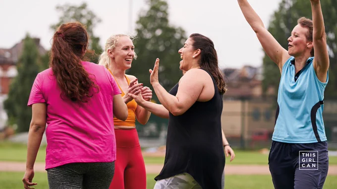 Four adult women joyfully interacting in a park, with one raising their arm in a celebratory gesture. They are wearing athletic wear. The logo "This Girl Can" is visible in the bottom right corner.