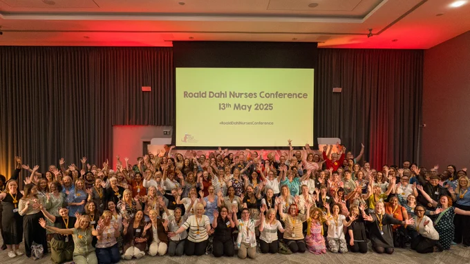 Group of 150 Roald Dahl Nurses smiling and waving at the camera in front of screen which reads 'Roald Dahl Nurses Conference 13th May 2025' 