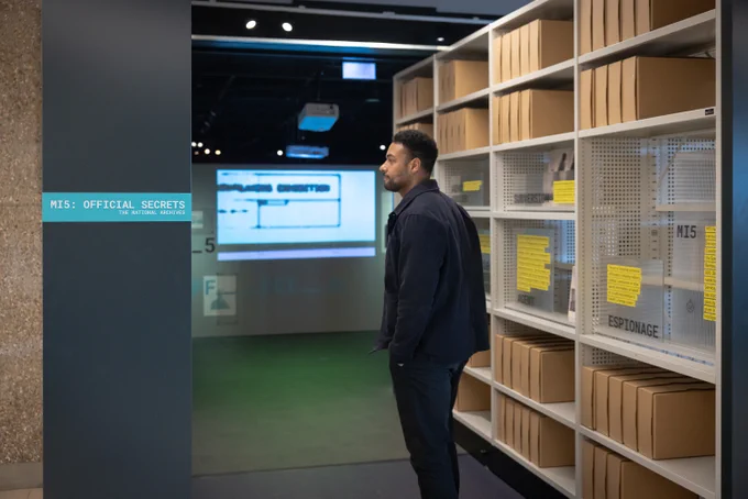 A man stand in the entrance of an exhibition titled 'MI5: Official Secrets', where the space is created out of mock archival shelving.