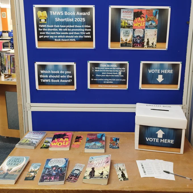 A blue display board shows the front covers of the shortlisted books and instructions on how to vote. In front is a wooden table with a ballot box, voting slips, and a copy of each of the books:
Angel of Grasmere by Tom Palmer
I Am Wolf by Alastair Chisholm
Impossible Creatures by Katherine Rundell
The Final Year by Matt Goodfellow
Shadowhall Academy: The Whispering Walls by Phil Hickes
When The Wild Calls by Nicola Penfold