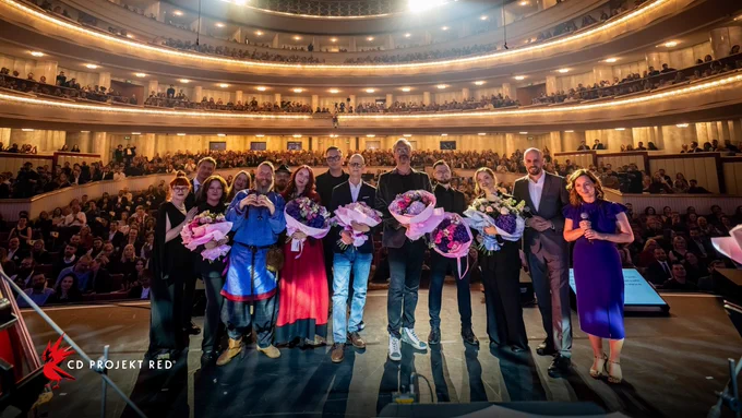 A group of CD Projekt Red developers stand with musicians at the front of a stage. Many of them hold flowers and smile. Behind them, hundreds of people sit in a round theatre.