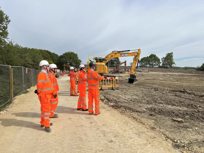 Group of Mining Remediation Authority and Kier colleagues dressed in orange high visibility overclothes in front of an area where topsoil has been stripped ready for construction and with a large digger and safety fencing in the background