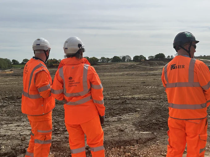 Three Mining Remediation Authority staff and a Kier colleague dressed in orange high visibility overclothes looking at an area of field where topsoil has been stripped ready for construction