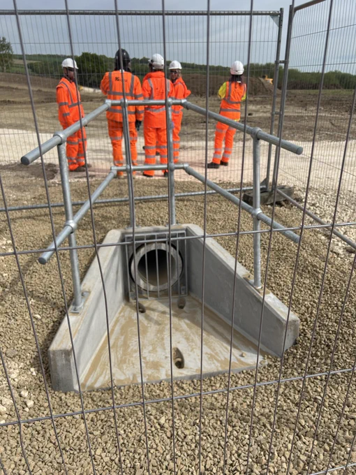 Group of Mining Remediation Authority and Kier colleagues dressed in orange high visibility overclothes in front of an area where topsoil has been stripped ready for construction. In the foreground there is a new concrete surface water drain surrounded by safety fencing