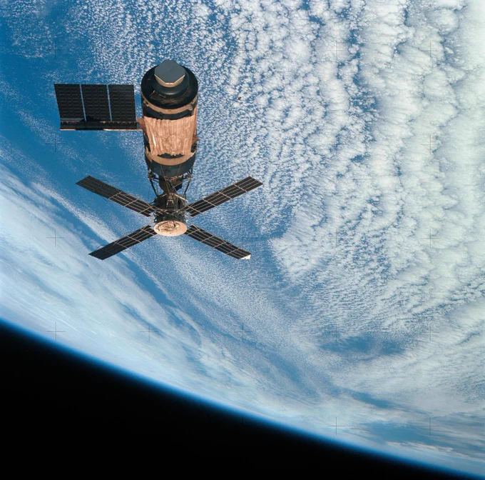 Skylab space station orbits above Earth, with a backdrop of textured clouds and the curve of the planet's horizon visible.