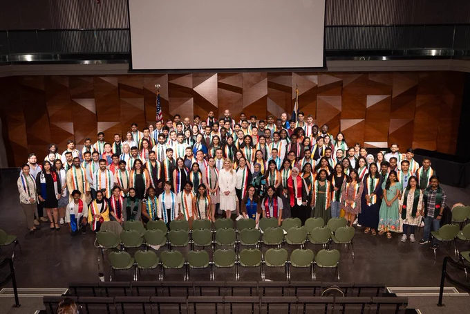 Large group of C S U international graduates pose on stage during the International Stole Ceremony. Graduates wear stoles representing their home countries, including colors and patterns from flags around the world. President Amy Parsons stands in the front row, centered among the graduates. An American flag and a Colorado state flag are visible in the background.